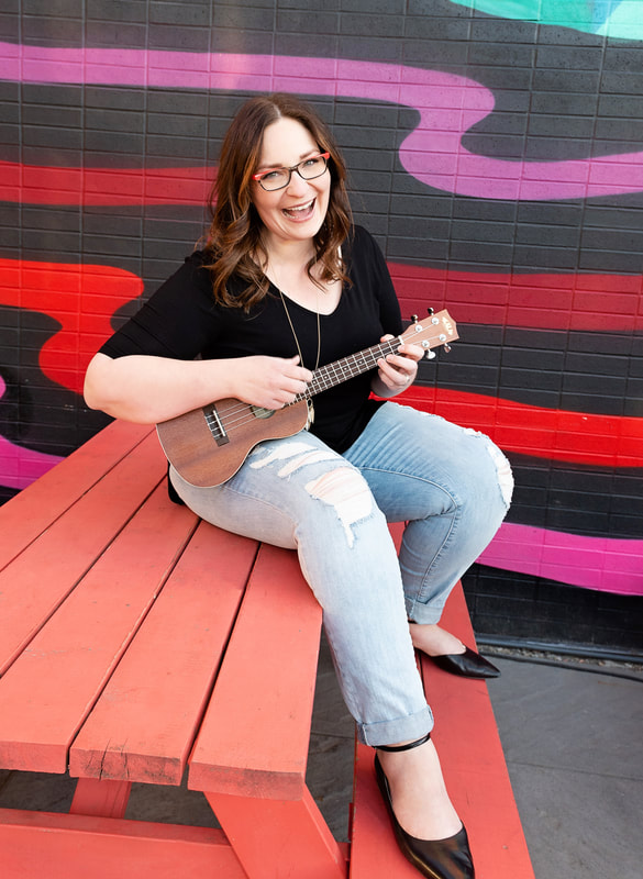 Photo of Beppie sitting on a picnic table, smiling, holding a ukulele. The brick wall behind her is painted black with purple, red and pink lines swirled horizontally.