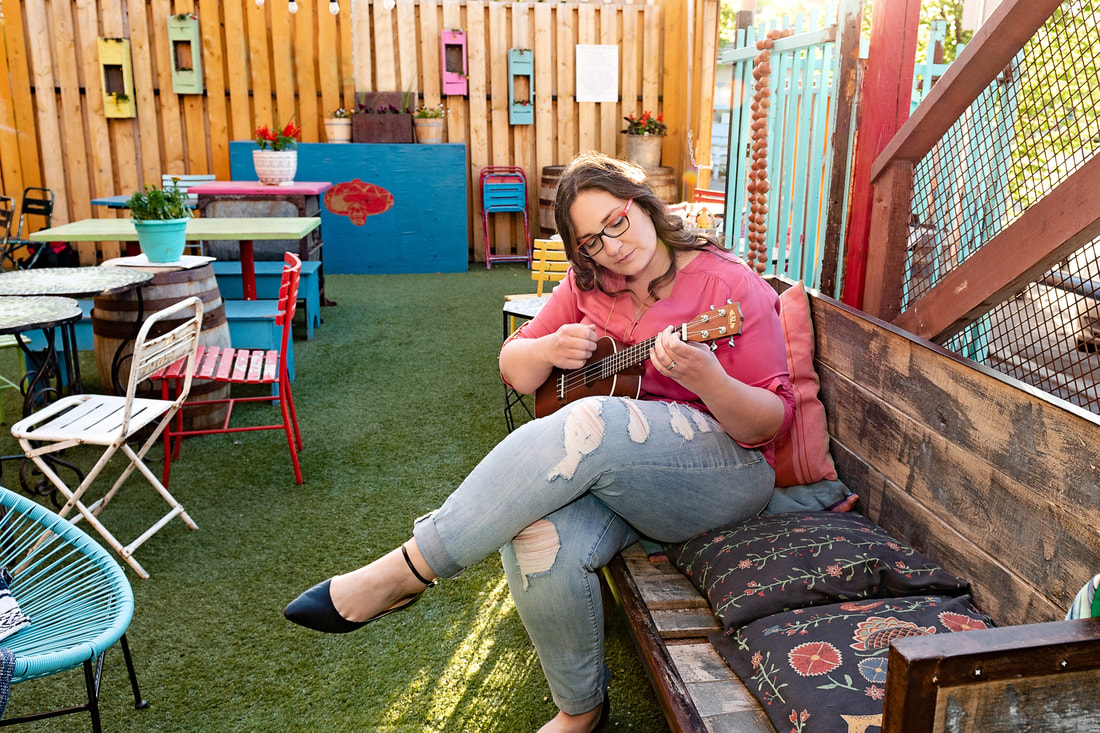 A photo of Beppie sitting on an outdoor patio playing a ukulele. She's wearing ripped jeans and a pink blouse.